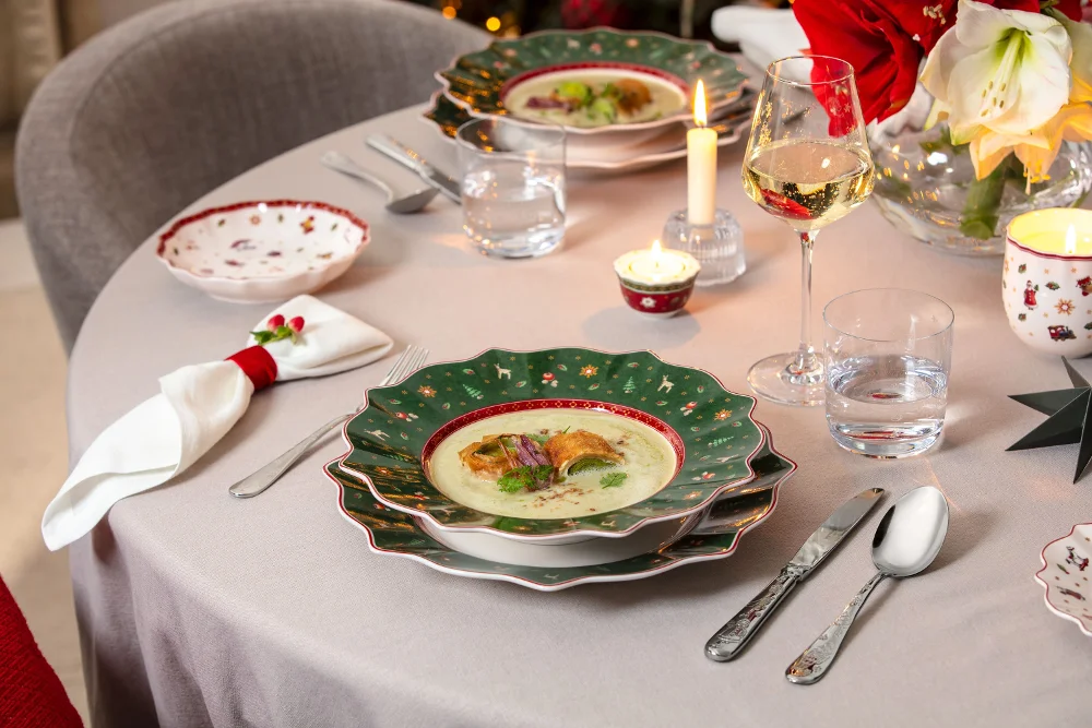  Una tavola imbandita a festa con una ciotola di zuppa, pane, posate, candele, un bicchiere di vino e decorazioni natalizie.