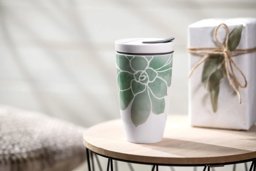 The Coffee ToGo mug from Villeroy & Boch, a ceramic travel mug with a green leaf design, stands on a wooden table next to a gift box tied together with string.