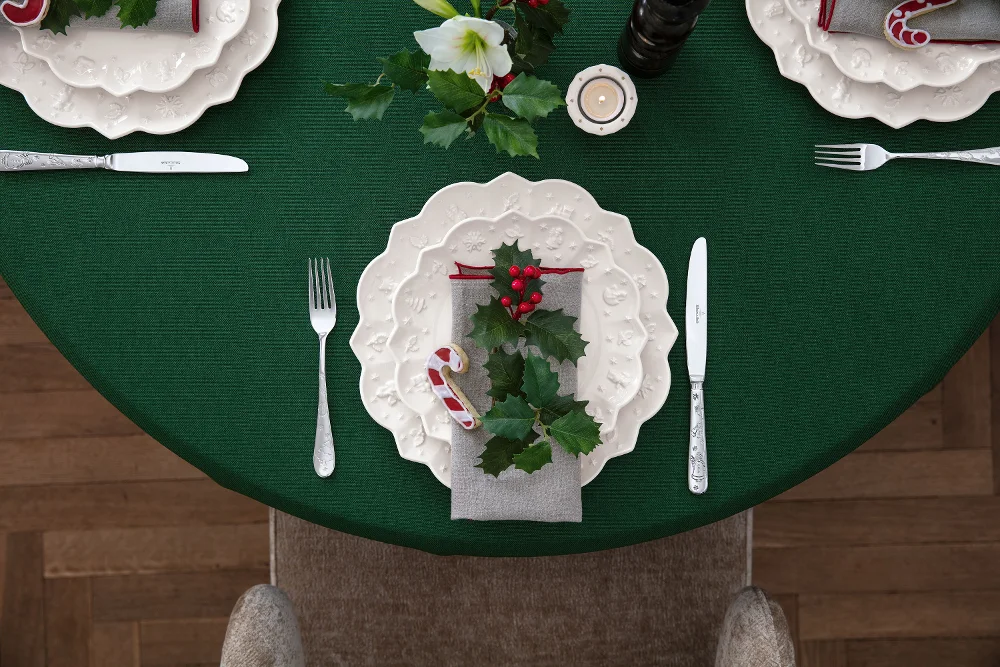 Una tavola apparecchiata per Natale con una tovaglia verde, piatti bianchi decorati con agrifoglio della collezione Toy's Delight Royal Classic di Villeroy & Boch, un bastoncino di zucchero, un rametto di agrifoglio sul tovagliolo e posate d'argento.
