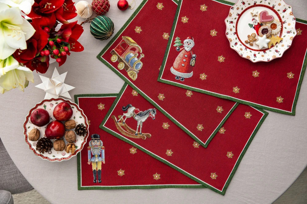 Toy's Delight Gobelin placemats with Christmas motifs Overhead view of four red Toy's Delight tapestry placemats featuring a nutcracker, rocking horse, train, and Santa Claus on a dining table, decorated with Christmas cookies, apples, walnuts, and festive ornaments