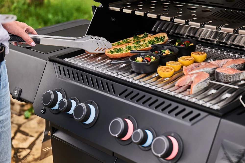 Eine Person grillt mit einer Zange Lachs, Zitronenhälften, Gemüse in kleinen Pfännchen und Brot auf einem Gasgrill im Freien.