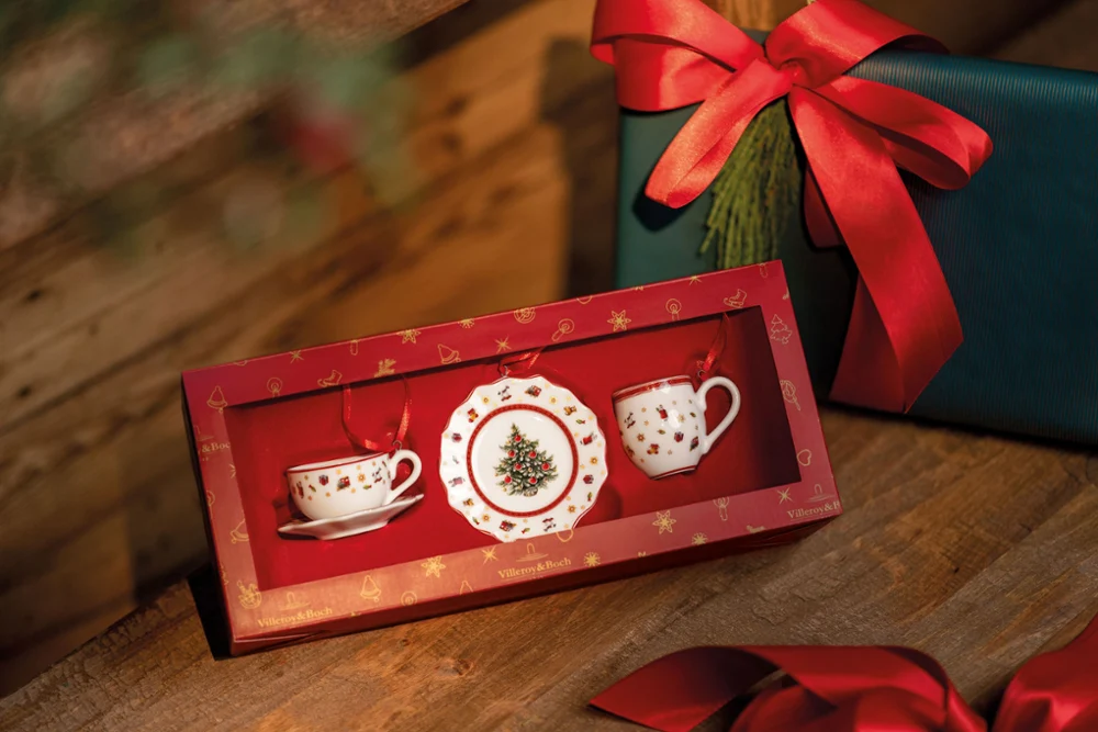 Toy’s Delight ornaments in a red box, consisting of a cup, saucer, and plate with Christmas tree and star motifs, are presented alongside wrapped presents with red ribbons.