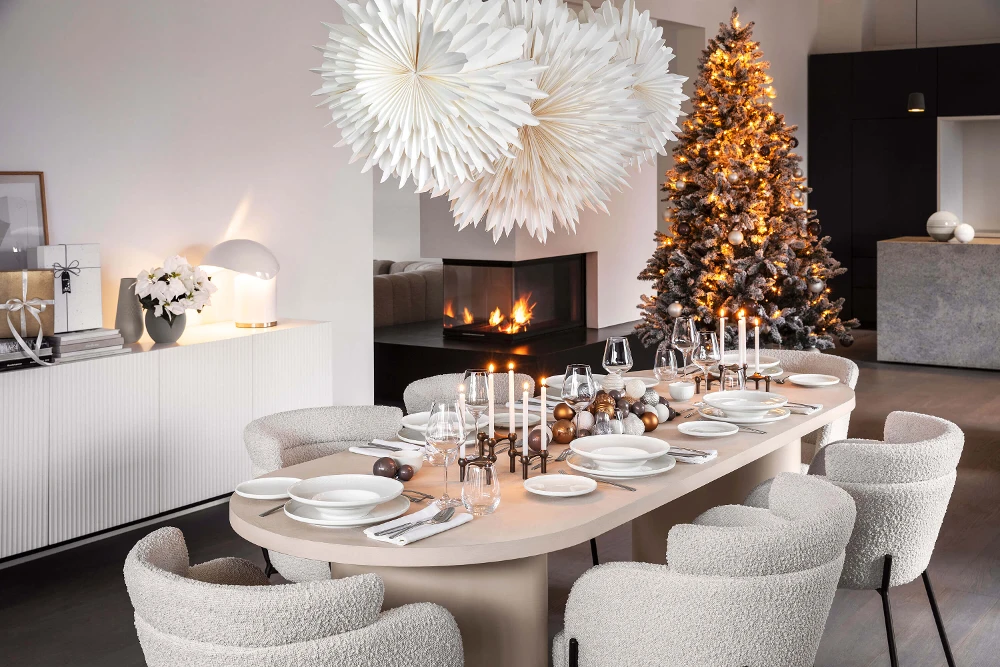 A modern dining room is set with white Afina plates and candles for a meal. There are also two large paper stars as decorations, a decorated Christmas tree, and a built-in fireplace.
