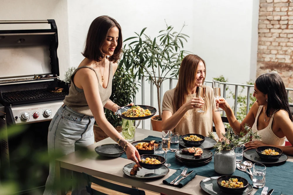 Summer BBQ with the Lave collection on a set table. Two friends enjoy summer delicacies on Lave plates and toast with a glass of sparkling wine, another person serves food.