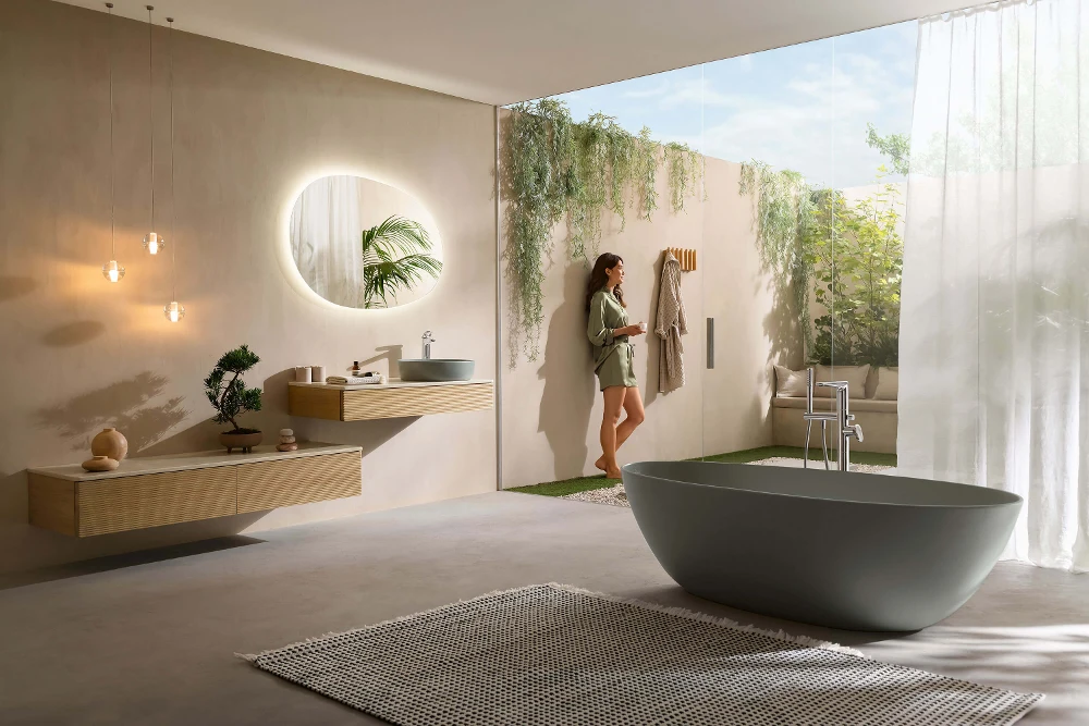 Modern bathroom with freestanding bathtub, countertop washbasin and a woman looking out at the greenery outside.