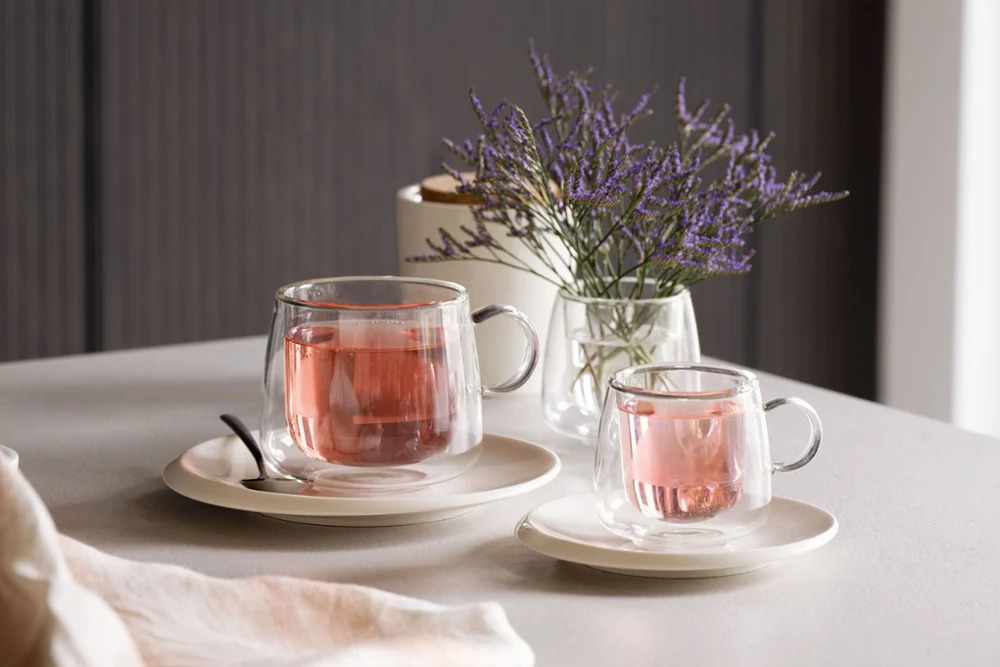 Artesano glass cups from Villeroy & Boch filled with pink tea, placed on saucers next to a spoon on a table that is decorated with a small vase of purple flowers.