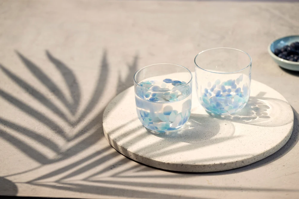 Two Like Aqua water glasses on a round tray Two Like Aqua water glasses with blue petals on a round tray, with palm leaf shadows on a light surface.