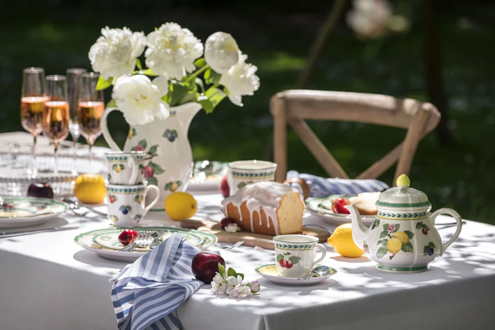 French Garden dinnerware on an outdoor table Outdoor table setting with flowered French Garden china, a teapot, rosé glasses, and a cake. White peonies in a vase and fruit are placed around the table.