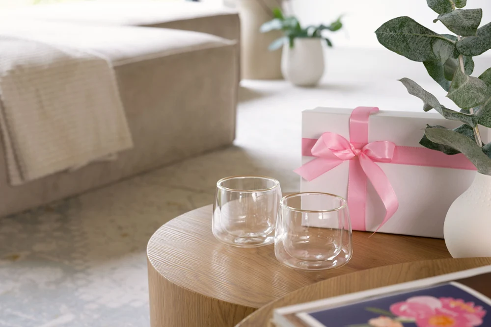 Two double-walled glasses with gold rims placed on a wooden table, next to a white gift box with a pink ribbon and a white vase with green eucalyptus leaves in a modern, cozy living room.