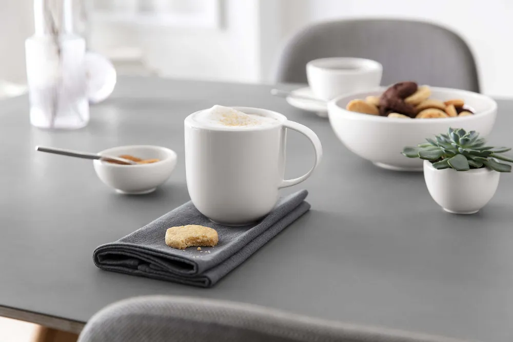 A mug of cappuccino on a gray napkin with a cookie, bowls of snacks, and a succulent on a modern dining table.