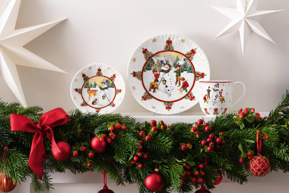 Toy's Fantasy Plates and Mug on a mantle Festive Christmas plates and a mug featuring snowmen are displayed on a mantle adorned with a garland of green pine, red ornaments, and a red bow. White star decorations are above.