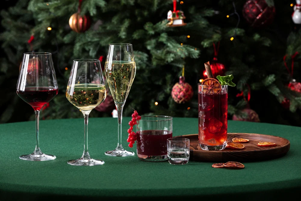 A green table with five different “Toy’s Delight” glasses from Villeroy & Boch, a wooden board with dried orange slices, and a decorated Christmas tree in the background.