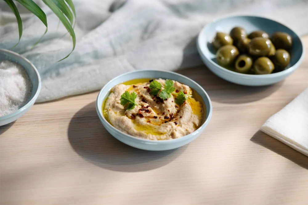 A Perlemor Aqua bowl with hummus stands next to a bowl of green olives on a light wooden surface. A Perlemor Aqua bowl with hummus, garnished with parsley and spices, stands next to a bowl of green olives on a light-colored wooden surface.