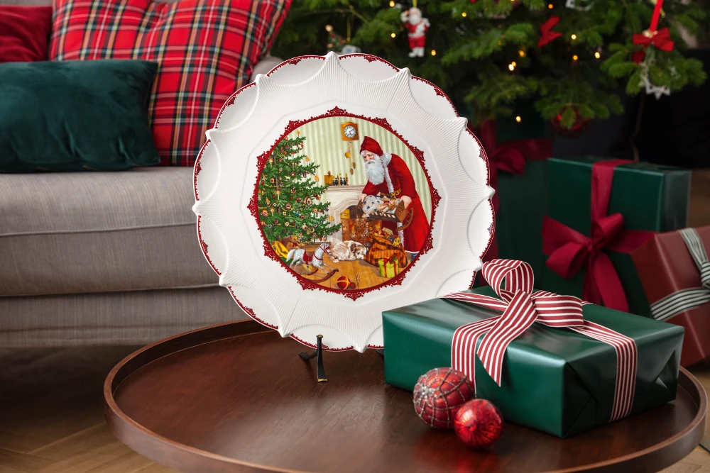 Toy's Fantasy large round platter and gift box A decorative plate depicting Santa by a Christmas tree, surrounded by gifts, on a wooden table with wrapped presents. Festive and cozy atmosphere.