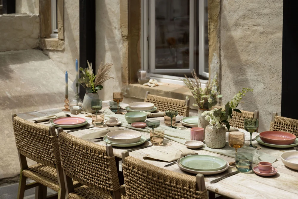 Rustic dining table with Perlemor dinnerware A rustic outdoor dining table set with woven chairs, pastel-colored Perlemor plates from like. by Villeroy & Boch, glasses and floral centerpieces against the backdrop of a stone wall with a window.