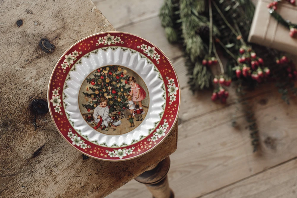 Toy's Fantasy Bowl on Wooden table with pine A decorative Christmas plate with a festive red border and floral designs depicts children decorating a Christmas tree. The setting is cozy and nostalgic.