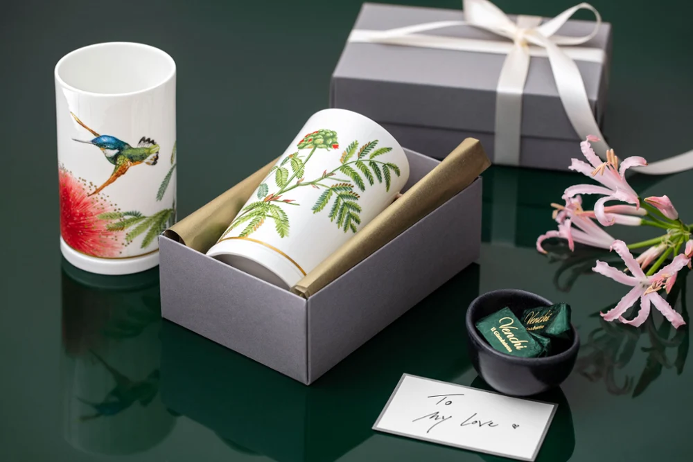 Presented are two Amazonia cups with flower and bird motifs, a gift box, a small bowl with wrapped items and a pink bow on a green background.
