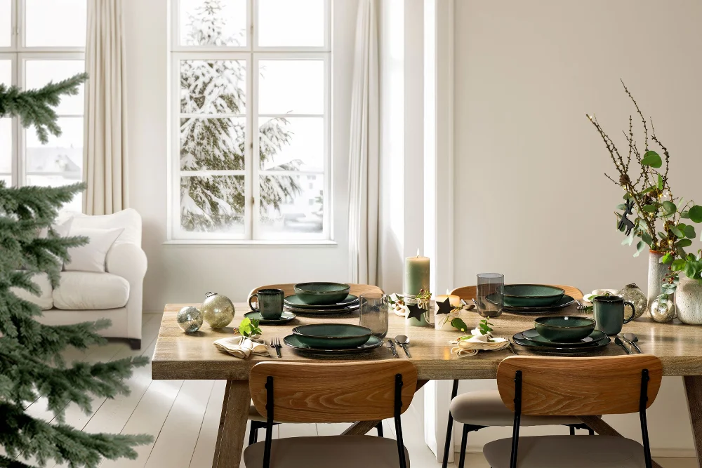 A bright and festive winter table setting featuring four place settings from Villeroy & Boch’s Lave Vert collection, including dinner plates, bowls, and handled mugs. The modern arrangement is complemented by a decorated Christmas tree and subtle seasonal decor, creating a serene holiday atmosphere with natural tones and handcrafted charm.