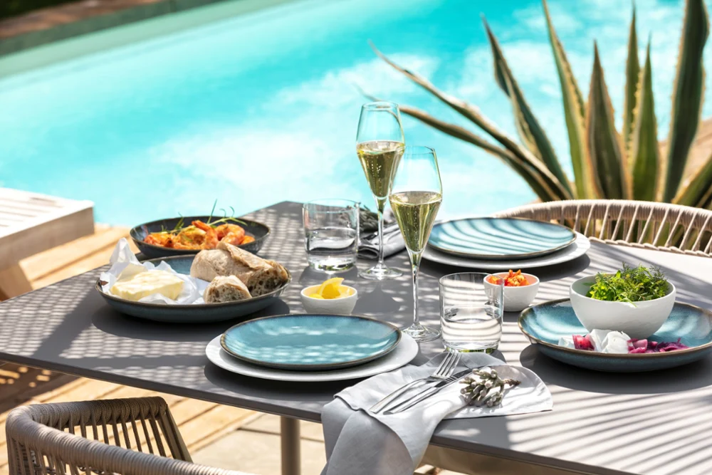 Al Fresco Poolside Table Setting A beautifully set outdoor dining table with plates, glasses of champagne, bread, and fresh salads beside a swimming pool.