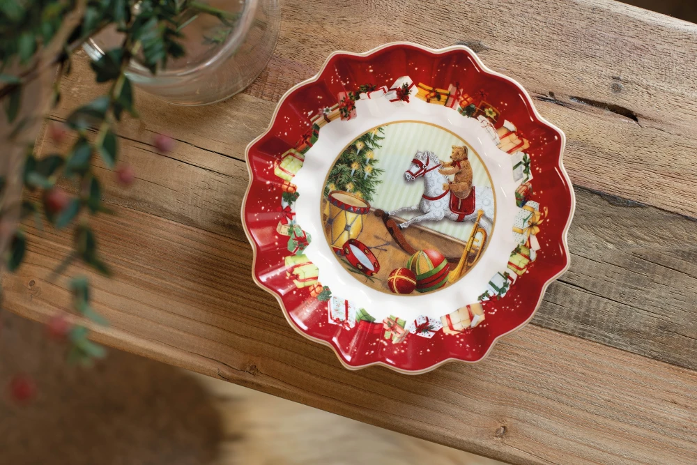 Toy's Fantasy round plate on wooden table Festive plate on a wooden table featuring a Christmas scene with a teddy bear on a rocking horse, surrounded by gifts and ornaments. The red border adds holiday cheer.