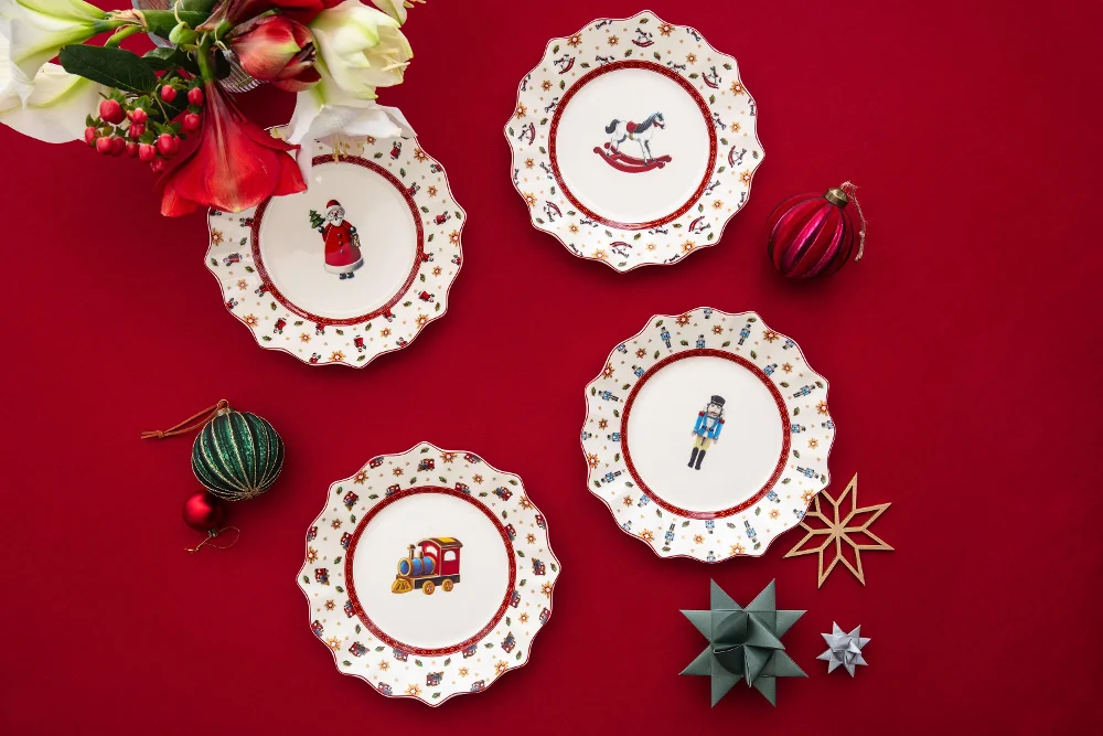 Quattro piatti natalizi decorativi con motivi festivi su una tovaglia rossa, circondati da ornamenti e fiori.