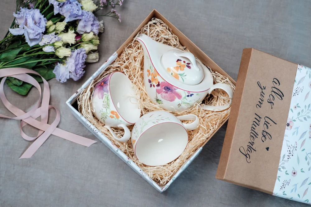A Mariefleur tea service in a gift box with straw filling, next to an open invitation card and purple flowers.