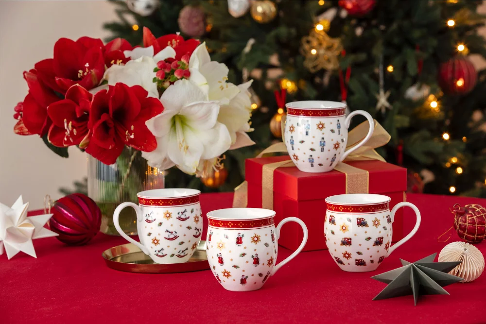 Quattro tazze festive su una tovaglia rossa con fiori, ornamenti, regali e un albero di Natale addobbato sullo sfondo.