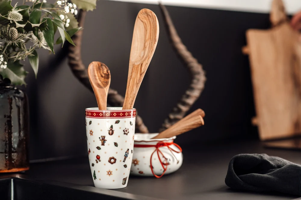 Two wooden cooking spoons in a Toy’s Delight To Go mug and a wooden ladle in a white bowl with a red rim, placed on a black kitchen counter with decorative elements in the background.