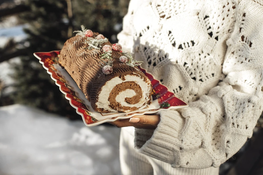 A woman holding a Toy's Fantasy Cake platter with cake A person in a white sweater holds a yule log cake on a festive plate outdoors. The cake is topped with sugared berries and leaves, evoking a cozy, winter feel.
