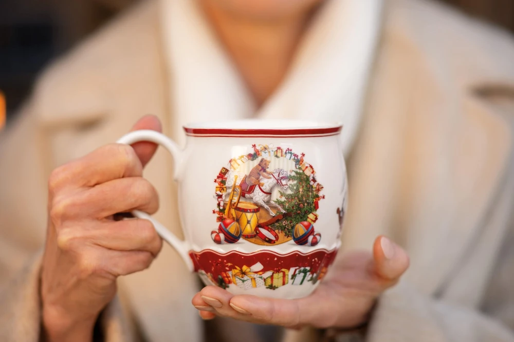A woman holding a Toy's Fantasy mug Smiling woman wearing a beige coat holds a festive mug with a Christmas design. The scene is warm and joyful, suggesting a holiday atmosphere.