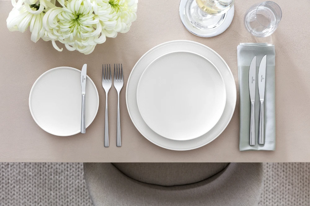 Table setting with Afina crockery and Piemont cutlery Sophisticated table setting with the Villeroy & Boch Piemont collection, including a white dinner plate, a salad plate, a cutlery set, glassware and a floral centrepiece on a beige tablecloth.