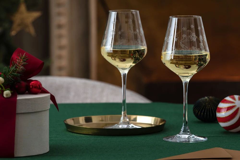 Two glasses of white wine on a green tablecloth are accompanied by a round white Toy’s Delight gift box from Villeroy & Boch with a red bow and festive decorations.