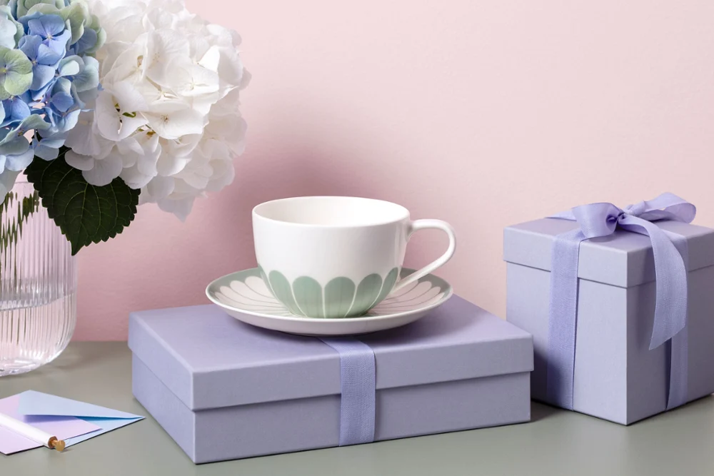 A Fleur teacup on a saucer with a green floral pattern stands on a lavender-colored gift box next to another gift box and a vase with white flowers.