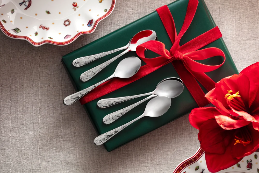 A green box with a red bow with five silver spoons, pictured on a gray background with a red flower and partially visible decorative plates from the Toy’s Delight collection by Villeroy & Boch.