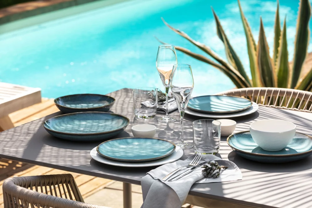 A table set with Lave and Manufacture plates A table set with Lave and Manufacture plates, glasses and flatware near a pool.