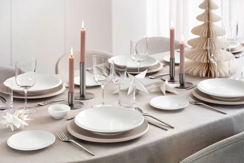 Top-down view of a modern, festive table setting featuring Villeroy & Boch’s NewMoon Beige collection: flat bowl, dinner and bread plates, paired with NewMoon cutlery, wine glasses and tumblers. Soft Christmas décor accents—such as light-toned ornaments, pine branches or candles—add a refined seasonal touch to the calm, natural tones and minimalist forms.