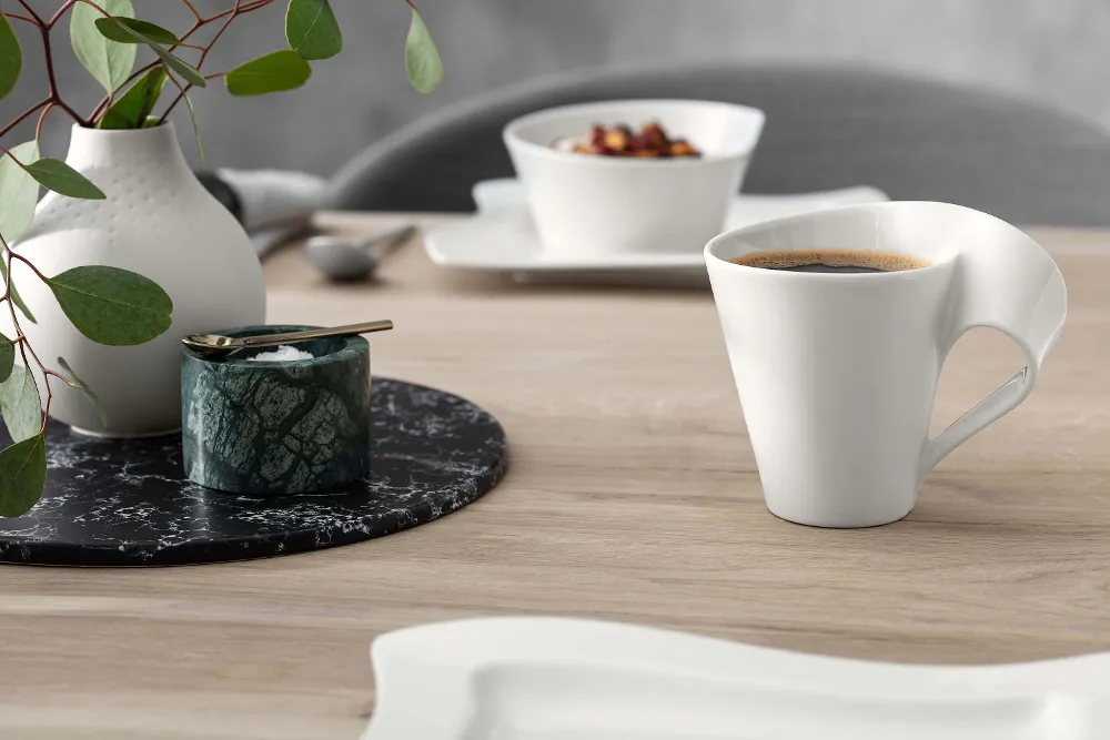 A modern white coffee mug with a unique handle design, placed next to a small jar and a vase with green leaves on a wooden table.