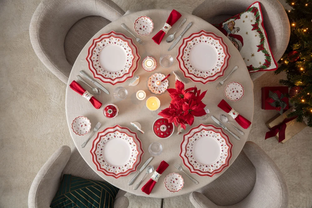 Festively decorated Christmas table with Toy's Delight tableware Top-down view of a round dining table elegantly set for Christmas with Villeroy & Boch's Toy's Delight porcelain plates, red napkins, candles, and a red poinsettia centerpiece next to a lit Christmas tree.