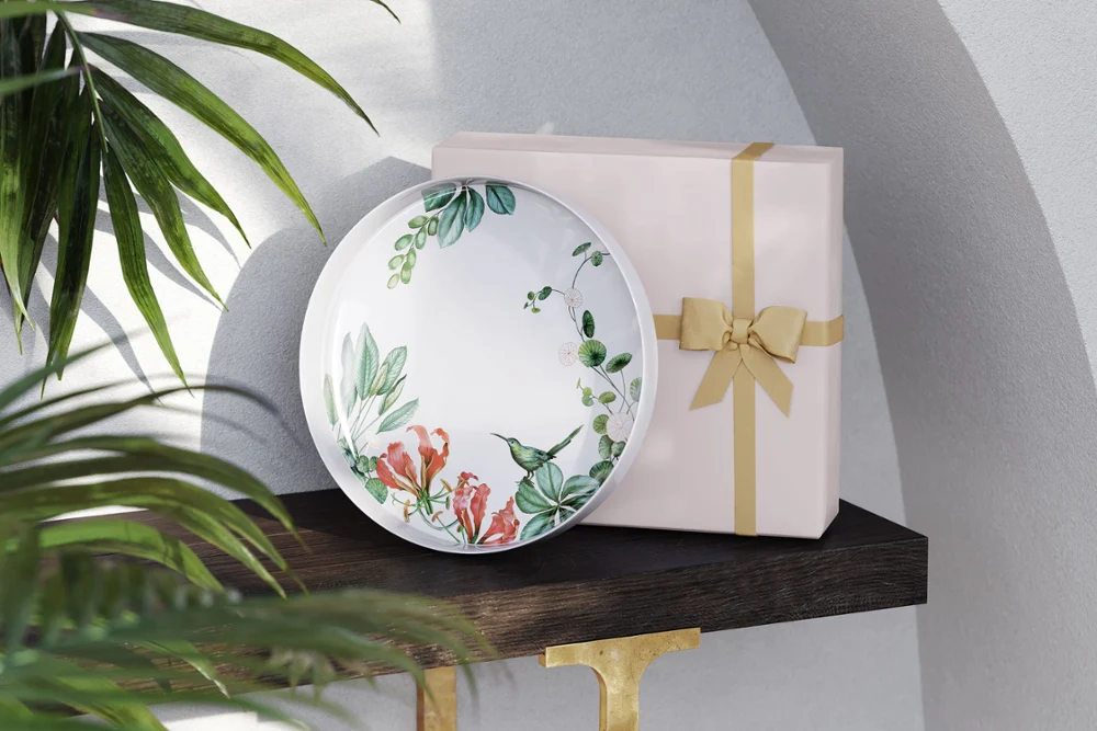 The Avarua plate by Villeroy & Boch stands next to a pink gift box with a yellow ribbon on a dark shelf.