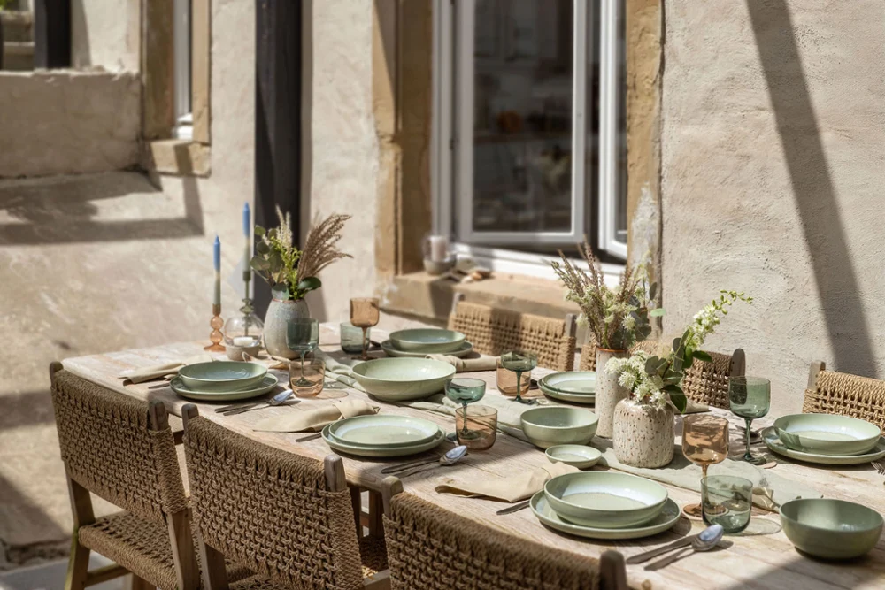A summery table setting with Perlemor A dining table set for six with green crockery stands outside next to a window and is decorated with flowers and candles.