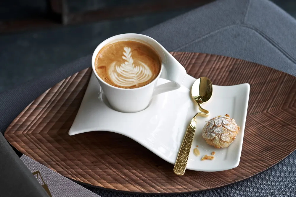 A Villeroy & Boch NewWave latte cup with latte art on a wavy white saucer is combined with a small cookie and a golden spoon, all beautifully arranged on a wooden tray.