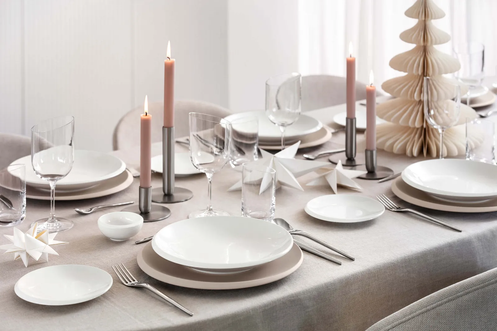 A beautifully set table with white NewMoon dinnerware, wine glasses, pink candles, silver flatware, and paper decorations on a beige tablecloth. There is also a small paper Christmas tree.