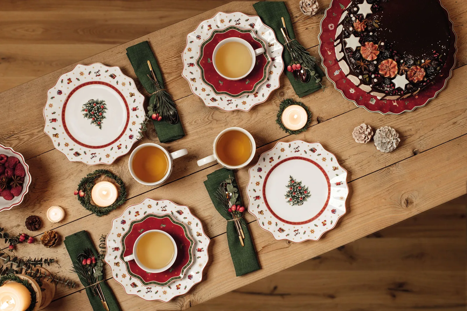 Festive holiday table setting with Toy’s Delight plates, cups of tea, a chocolate cake, and candles on a wooden table.