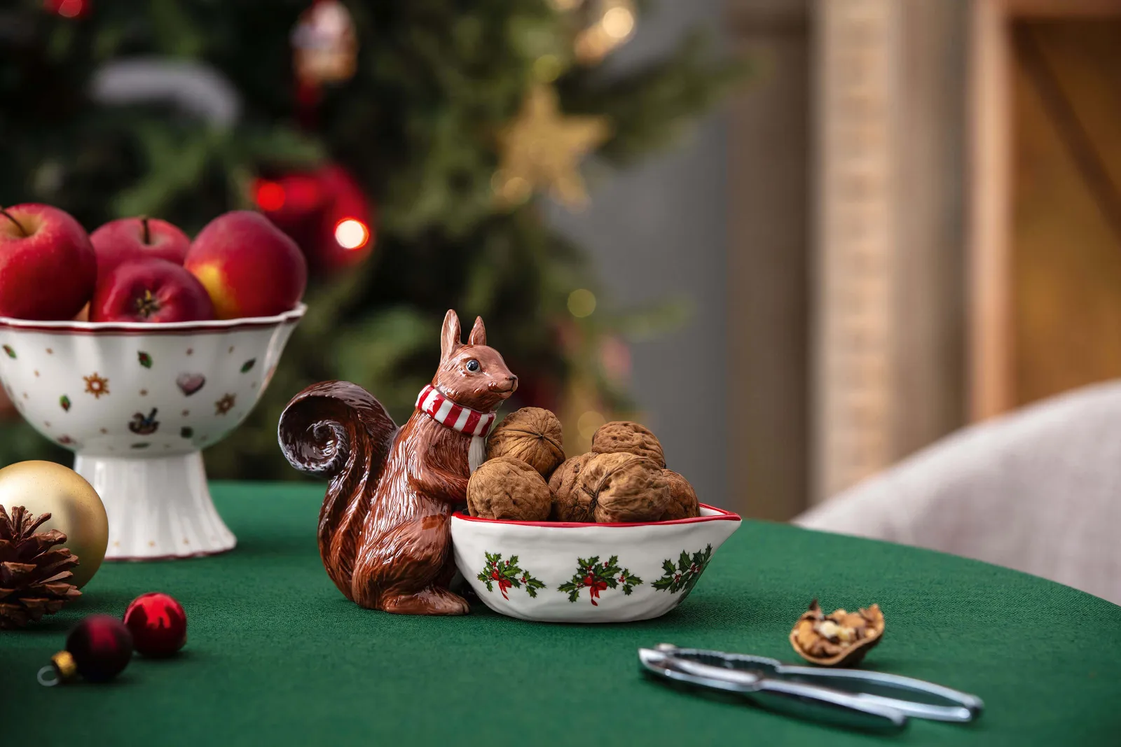 A bowl from Toy’s Fantasy decorated with a squirrel figure contains walnuts next to a nutcracker on a green tablecloth. Next to it is a bowl of red apples, with a decorated Christmas tree in the background.