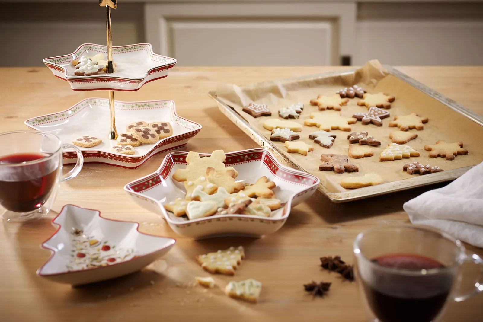 A wooden table with star-shaped bowls from Villeroy & Boch Winter Bakery Delight, on which decorated cookies are placed, and a baking tray with festive cookies on baking paper. There are two glasses with a red beverage on the table.