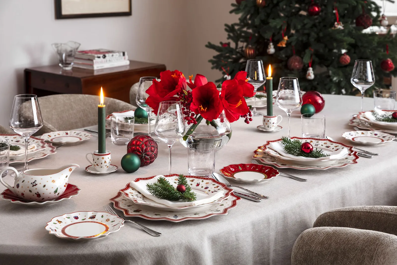 A festive dining table setting with elegant Toy’s Delight dinnerware, burning candles, red flowers in a vase, and Christmas decorations, with a decorated Christmas tree in the background.