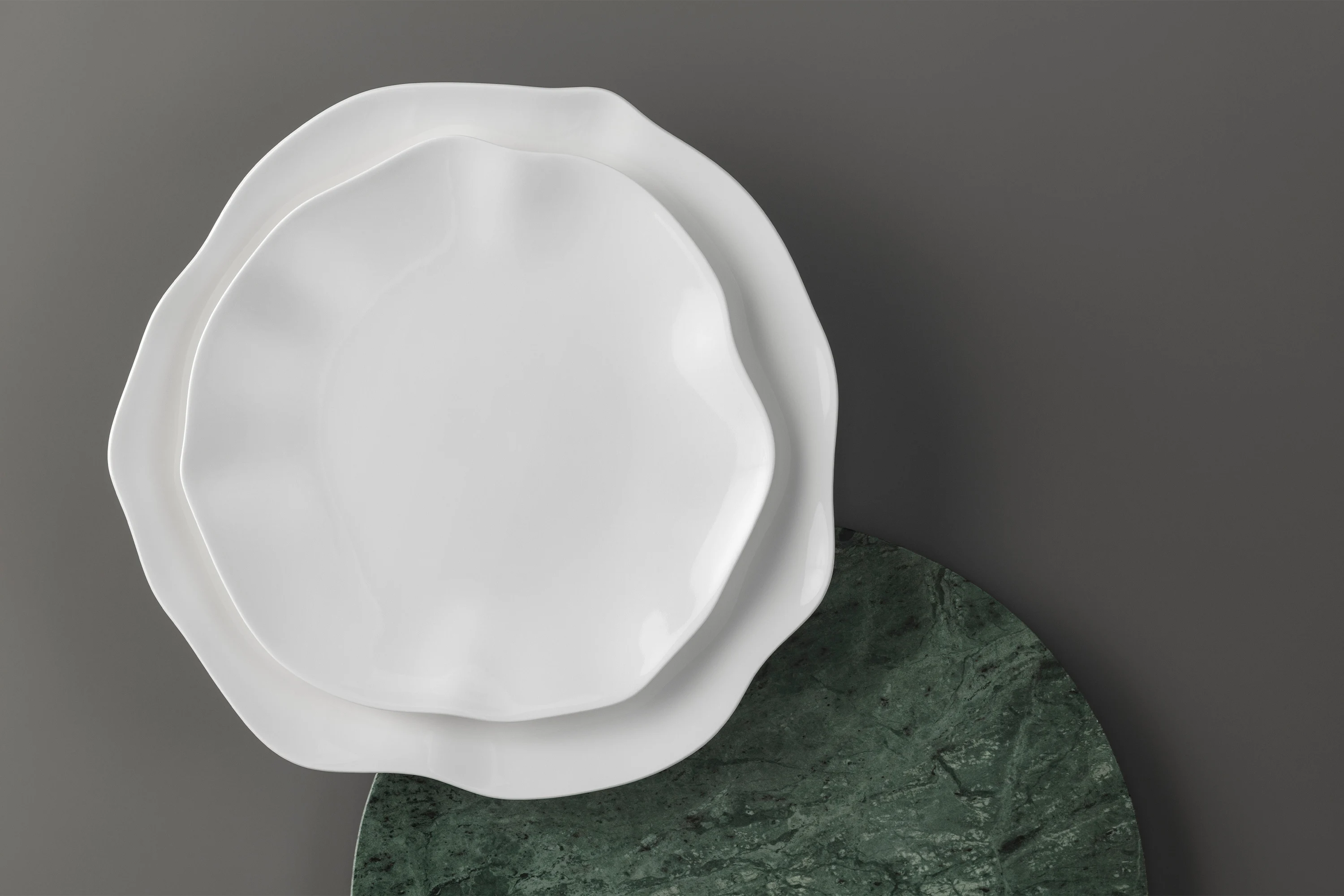 Deux assiettes Pura blanches de forme irrégulière sont empilées sur une assiette ronde vert foncé devant un fond gris.