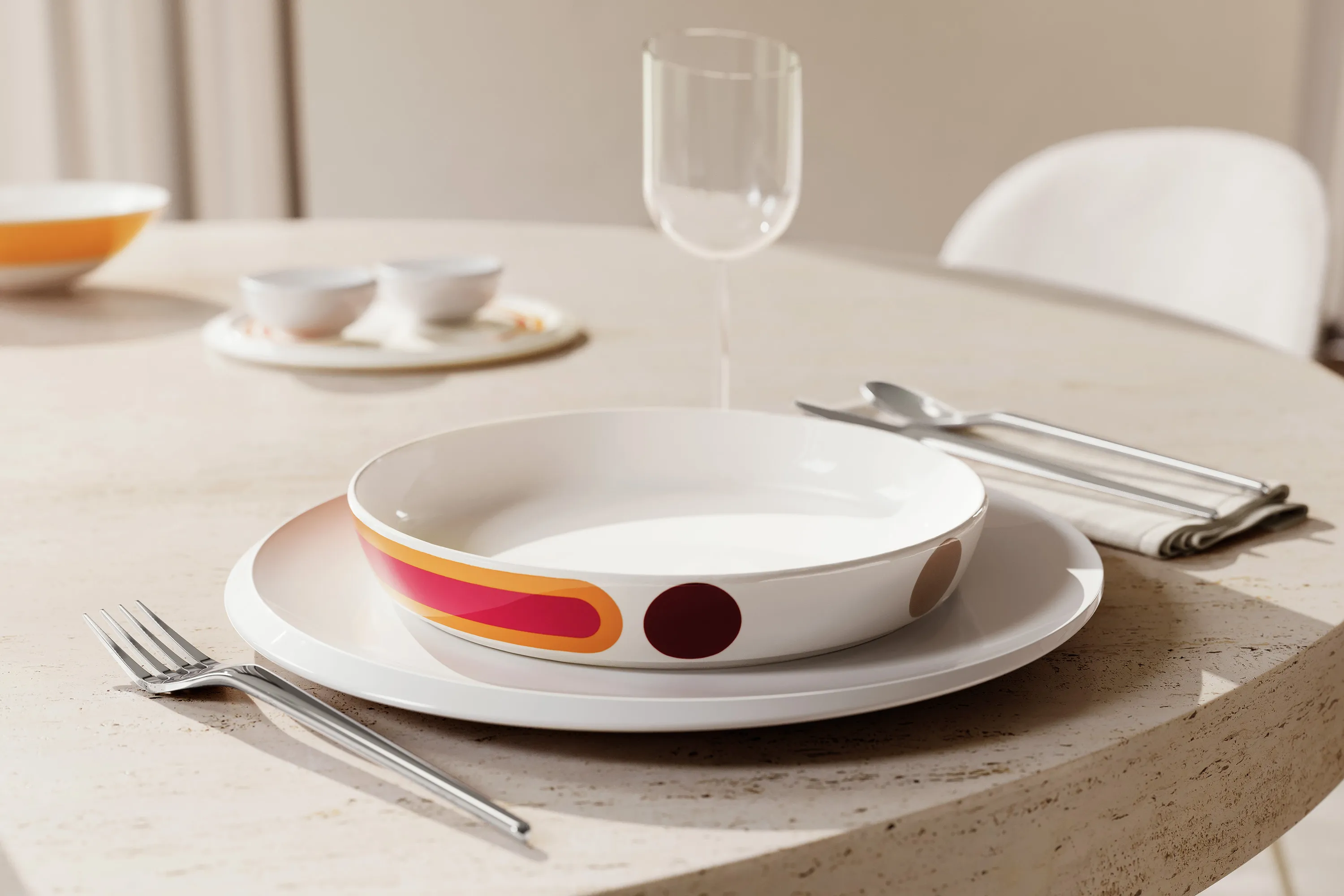 A table setting with a patterned La Boule bowl on a New Moon plate, fork, knife, spoon, and wine glass on a light-colored round table.