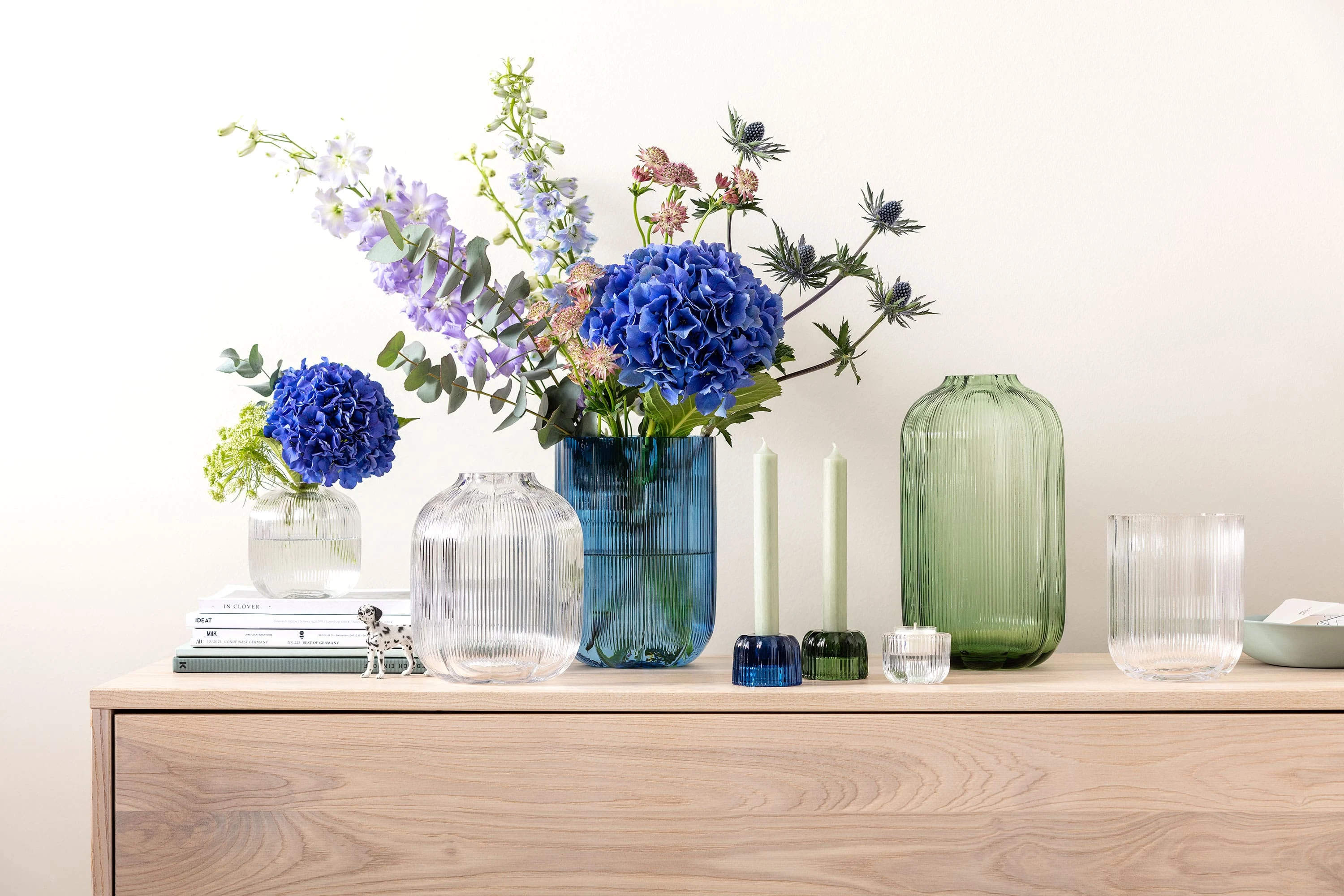 A sideboard with Fleur vases from Villeroy & Boch, decorated with blue and purple flowers, next to two green candles and stacked books.