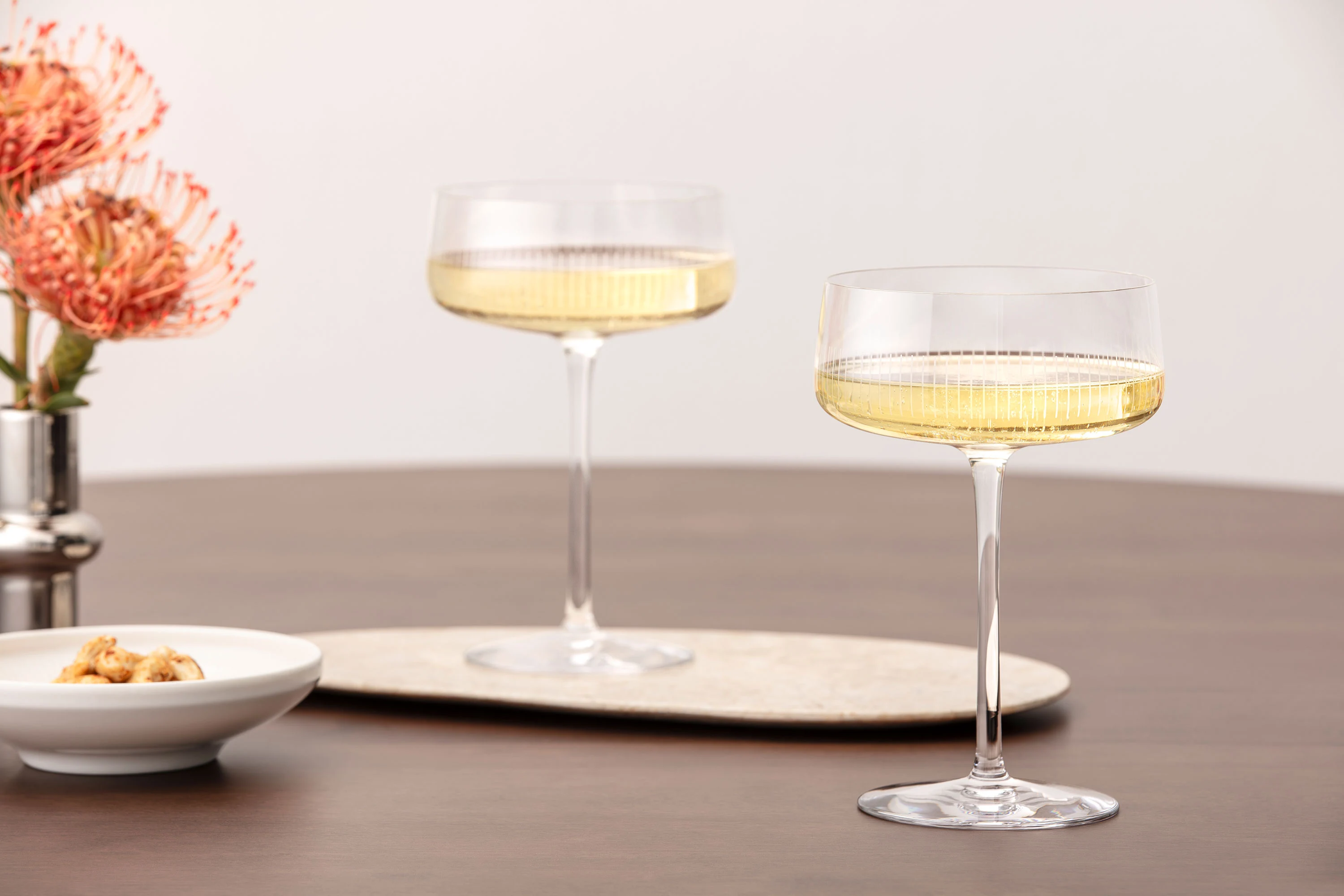 Two Afina glasses filled with sparkling wine on a wooden table, next to flowers and a small bowl.